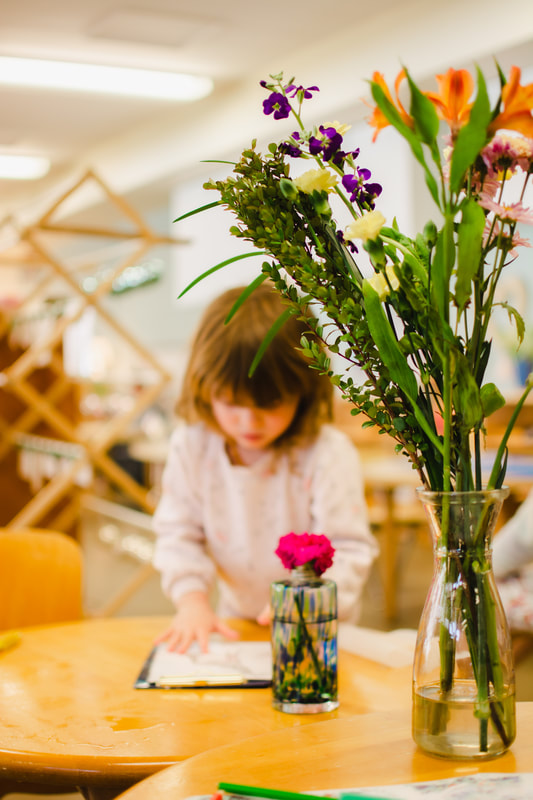 A vase of flowers sits on a table in a classroom with a child working in the background.
