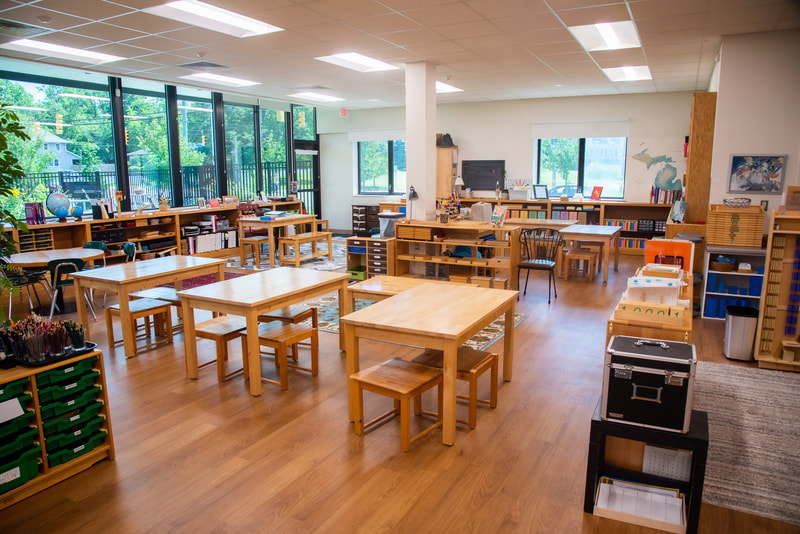 A bright and clean classroom showing many tables and chairs. The left wall is open with all windows while the back all has two windows. Shelves with works are all along the edges of the classroom layout.