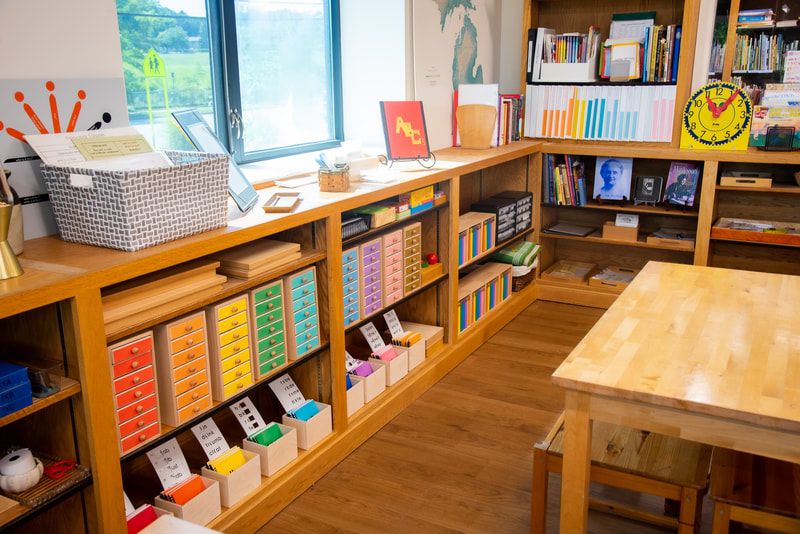 Low, child-sized corner shelves featuring color coordinated language works in wooden activity boxes as well as books and a photo of Maria Montessori. Next to the shelves is a child-sized table. Behind the shelves are more windows.