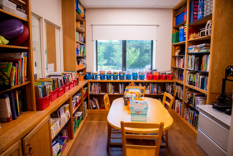The classroom library is shown with a table and chairs in the center surrounded by well organized shelves and baskets with various resources and materials.