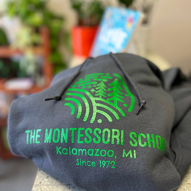 The Montessori School green logo sweatshirt, charcoal color is sitting on top of the 750 front desk.