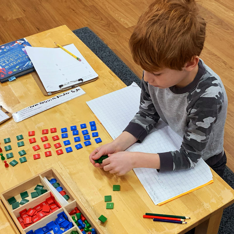 Looking down on to a student with red hair and a black camouflage shirt who is sitting on the floor and just starting to lay down their work tiles in color groups. They have paper below their elbows that are resting on the table.