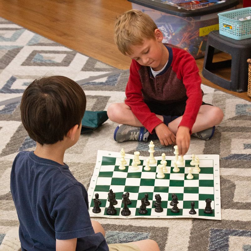 Two students are shown sitting on the floor about to play chess. One student sits facing the camera and debating their first move of Chess. The other student is facing away from the camera as they focus on their opponent.