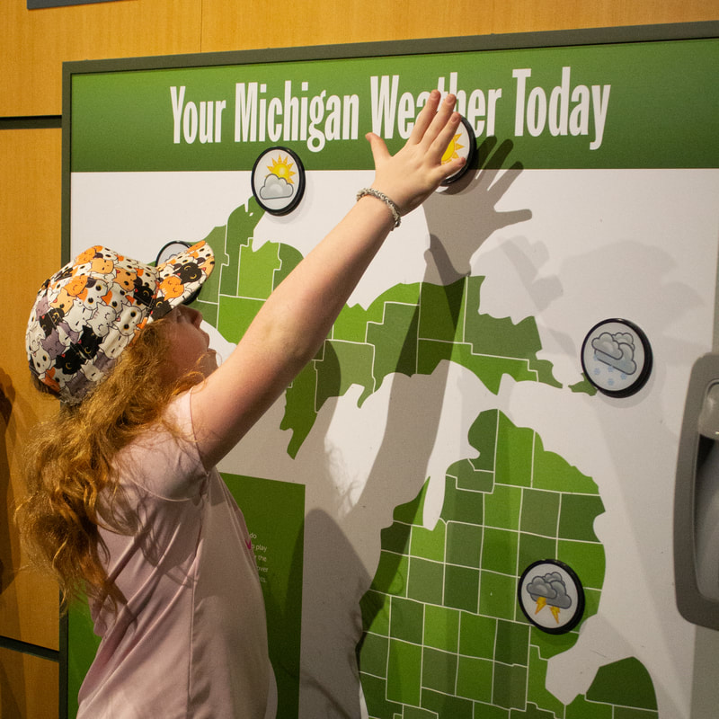 Child wearing a hat covered in cats and a pink shirt stretches their arm up to the words, "Your Michigan Wether Today". They are standing in front of a weather map of Michigan that features movable weather magnets for play.