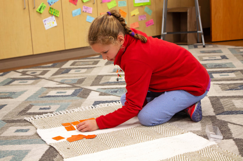 Student is on a mat and just beginning to lay out bright orange cards for culture. They are wearing a bright red sweatshirt and light blue pants.