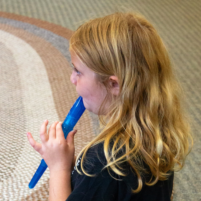 Side profile of student shown in an open classroom as they focus to the left of the image and are playing a vibrant blue, plastic recorder. Child has blond hair and a black shirt.