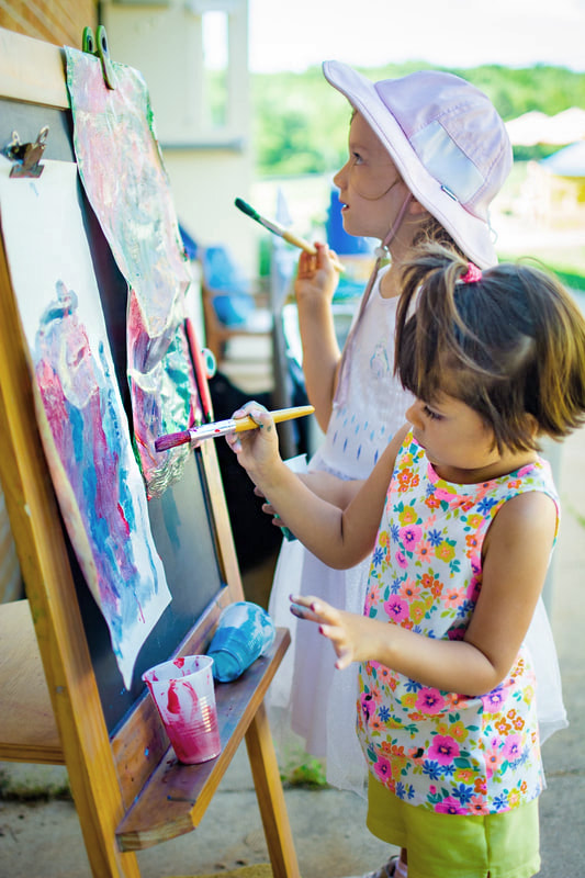 Two students standing outside under the shade of the building and using easels to paint. The student in front has bright flowers in pinks, yellows, and blues while one hand outstretched as they focus. The other student is determining what to do next on the painting and is dress with a hat and light pink dress.
