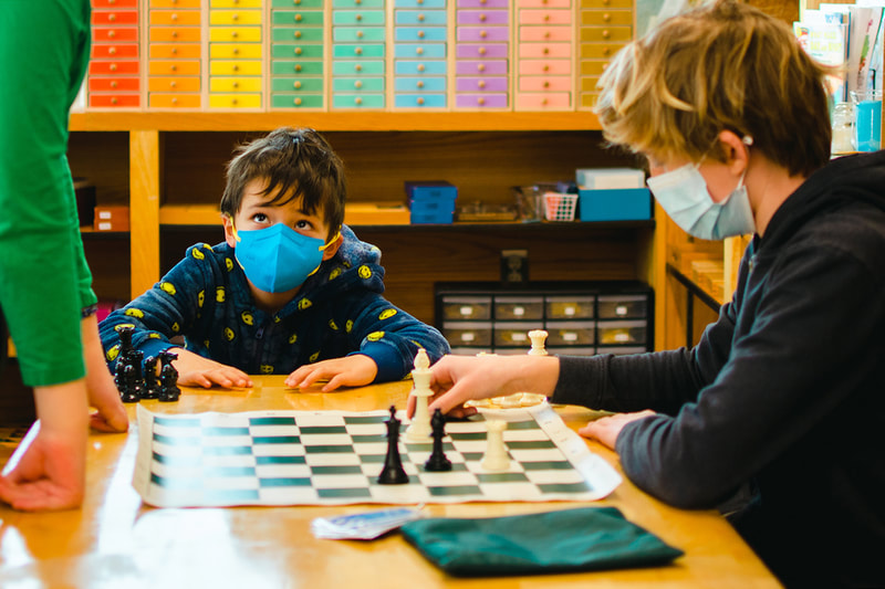 One child eagerly looks on as two older children play a game of chess
