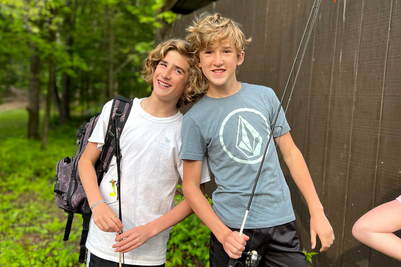 Two students lean their shoulders closer to one another as they pose for a photo with fishing poles, white and grey t-shirts, and a backpack. They are standing next to a small shed in the woods.