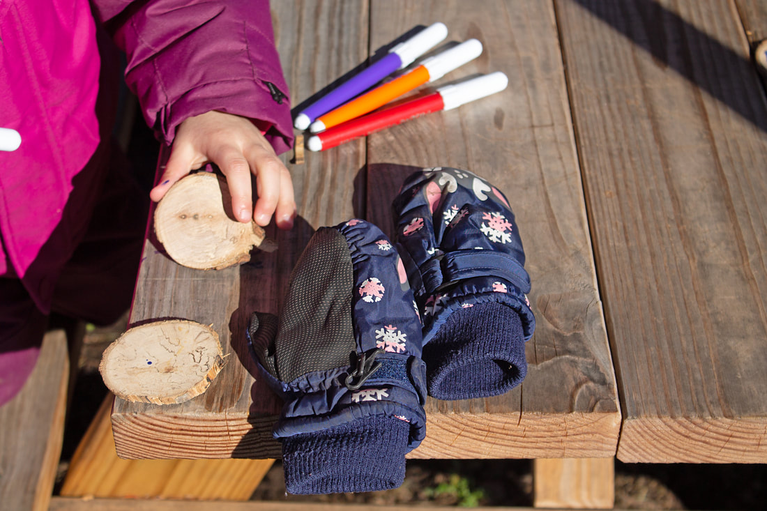 Hand seen holding a wooden circle for decoration; next to the hand are three markers (purple, orange, and red). Dark blue gloves sit in the middle of the image at the end of a picnic table. The child is wearing a thick winter coat.