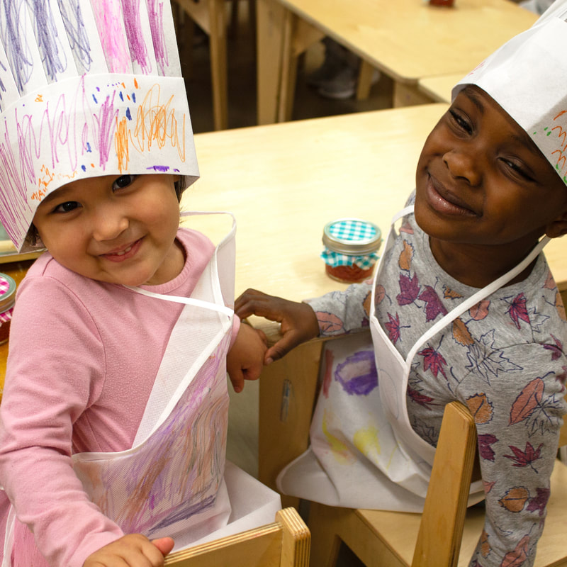 Two small Culinary Art Week campers are shown in paper chef hats and aprons they personally decorated. They are seated and turned away from their table; behind them is a jar of strawberry jam with a plaid fabric decor on top.