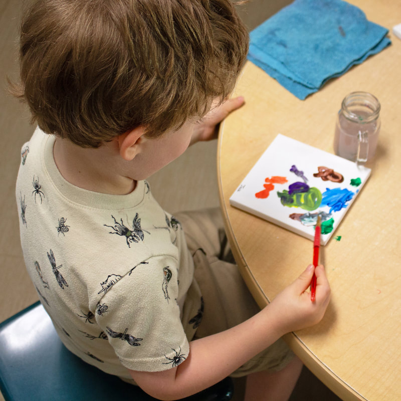 Child is paining on a small canvas using one of every color and creating small patches of each. They are facing the circle table they are using for their workspace with a blue rage and jar of water to the top and left of their cavas. The child is wearing a light tan shirt is various black illustrations of bugs.