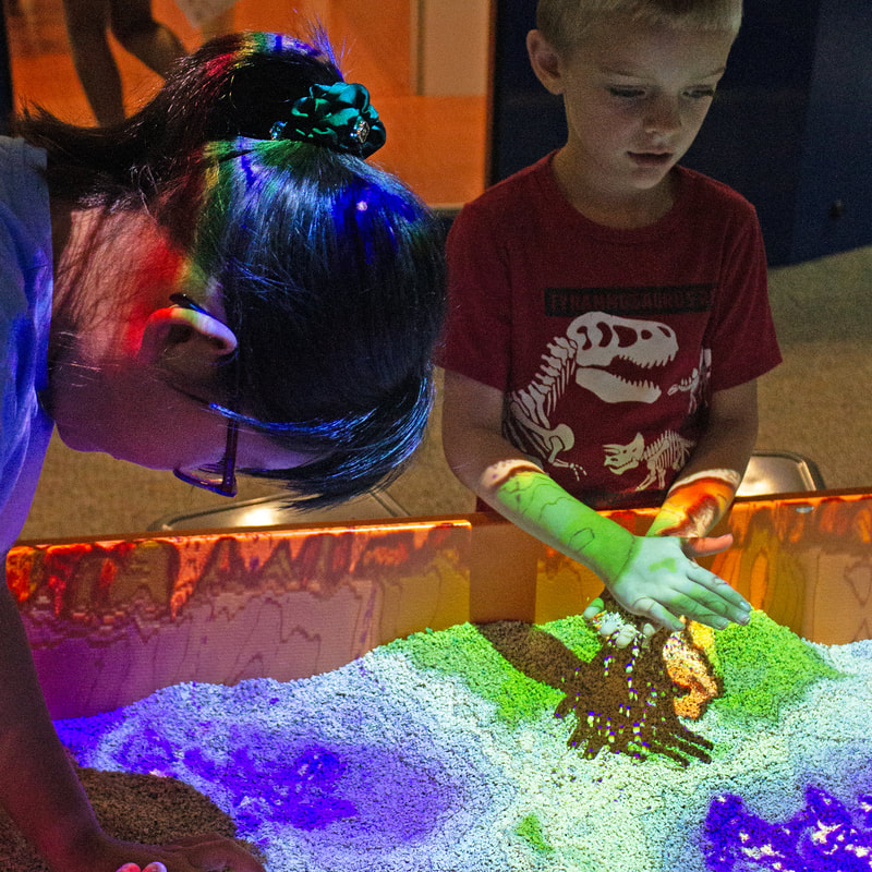 Two students are using a sand display at Kalamazoo Valley Museum where the light creates a map showing the different depths of the sand. The student to the left of the image is facing directly down with their hair up and glasses. The student in the back ground of the image has sand falling from their hands and is looking off to the right at a separate part of the display while wearing a bright red dinosaur shirt.